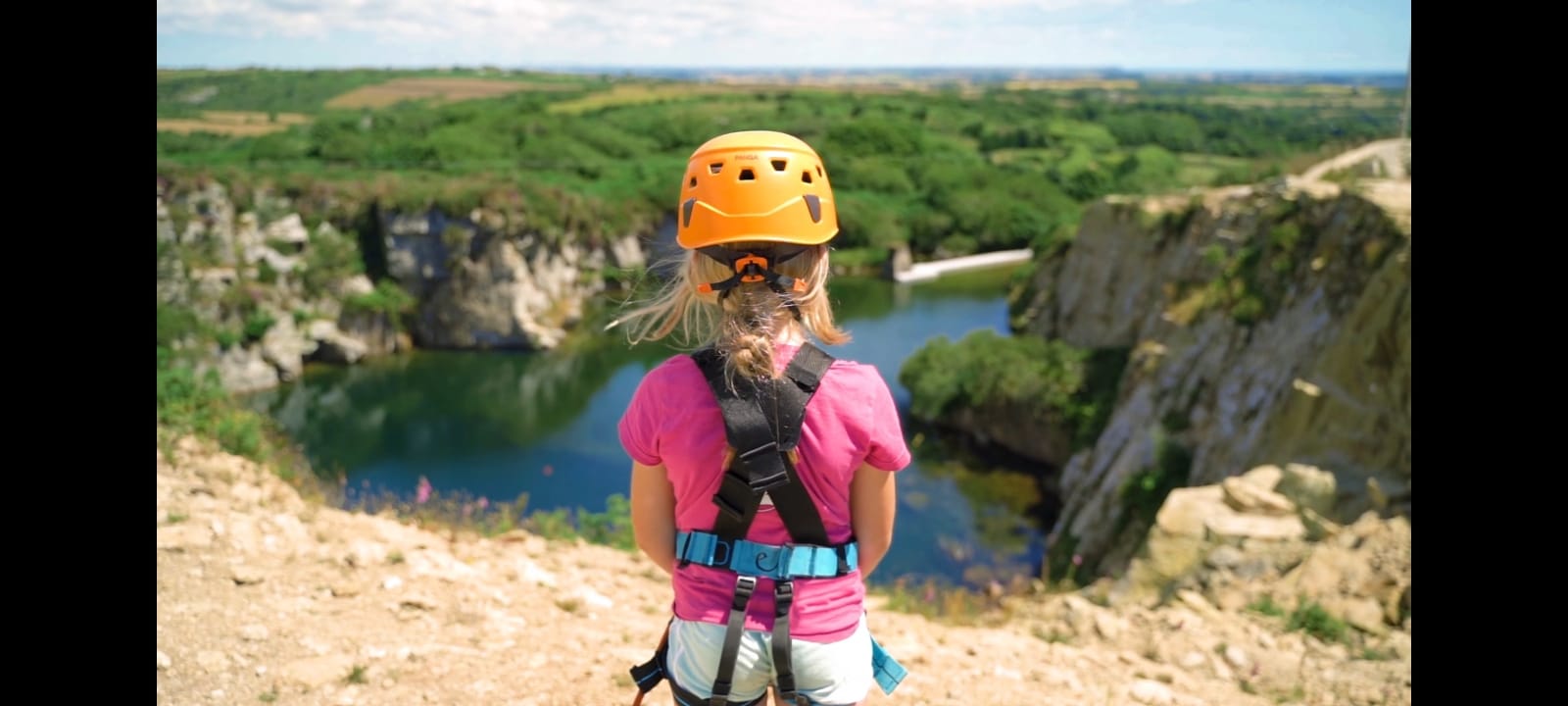Photo of young girl in a pink tshirt over looking a britsh quarry. She is hearding a orange hard hat and safety harness